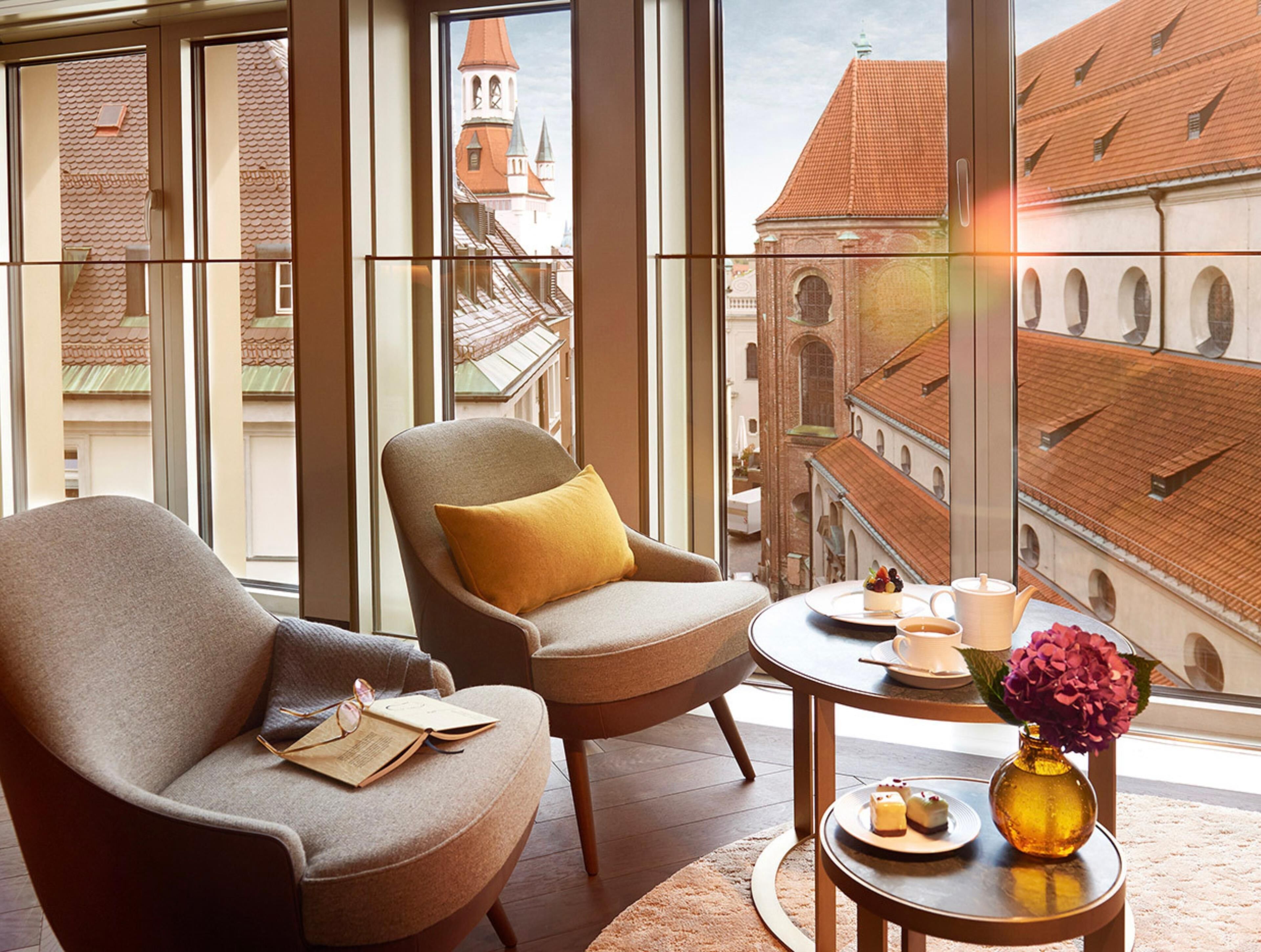 Quarto com duas poltronas, uma mesa redonda pequena com um vaso e doces, e vista para edifícios históricos por grandes janelas.