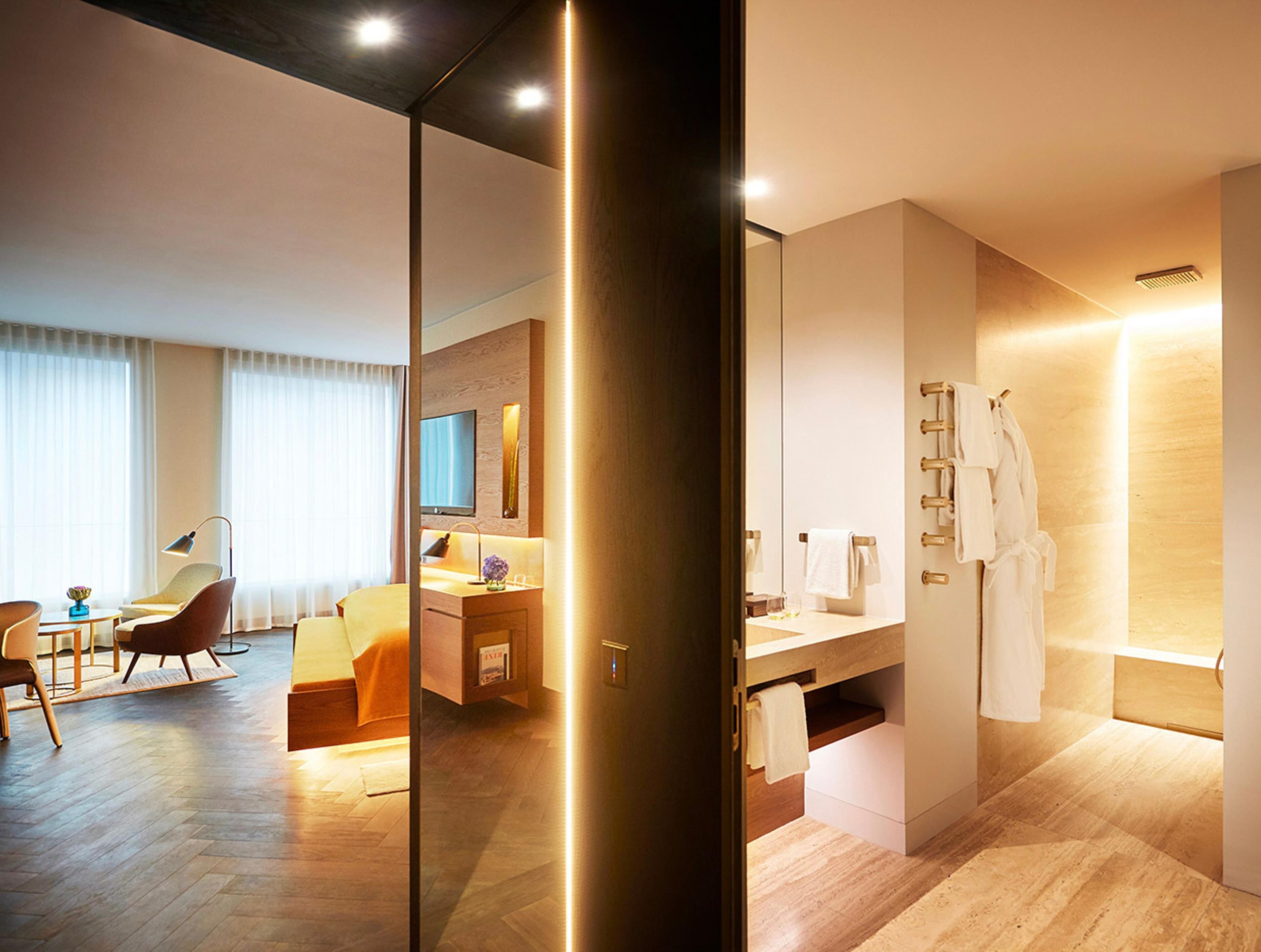 A modern hotel room with a wooden floor, a sitting area with a chair and a table, a large mirror, a bathroom with a bathtub and a towel rack.