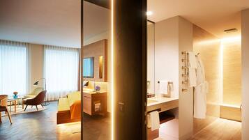 A modern hotel room with a wooden floor, a sitting area with a chair and a table, a large mirror, a bathroom with a bathtub and a towel rack.