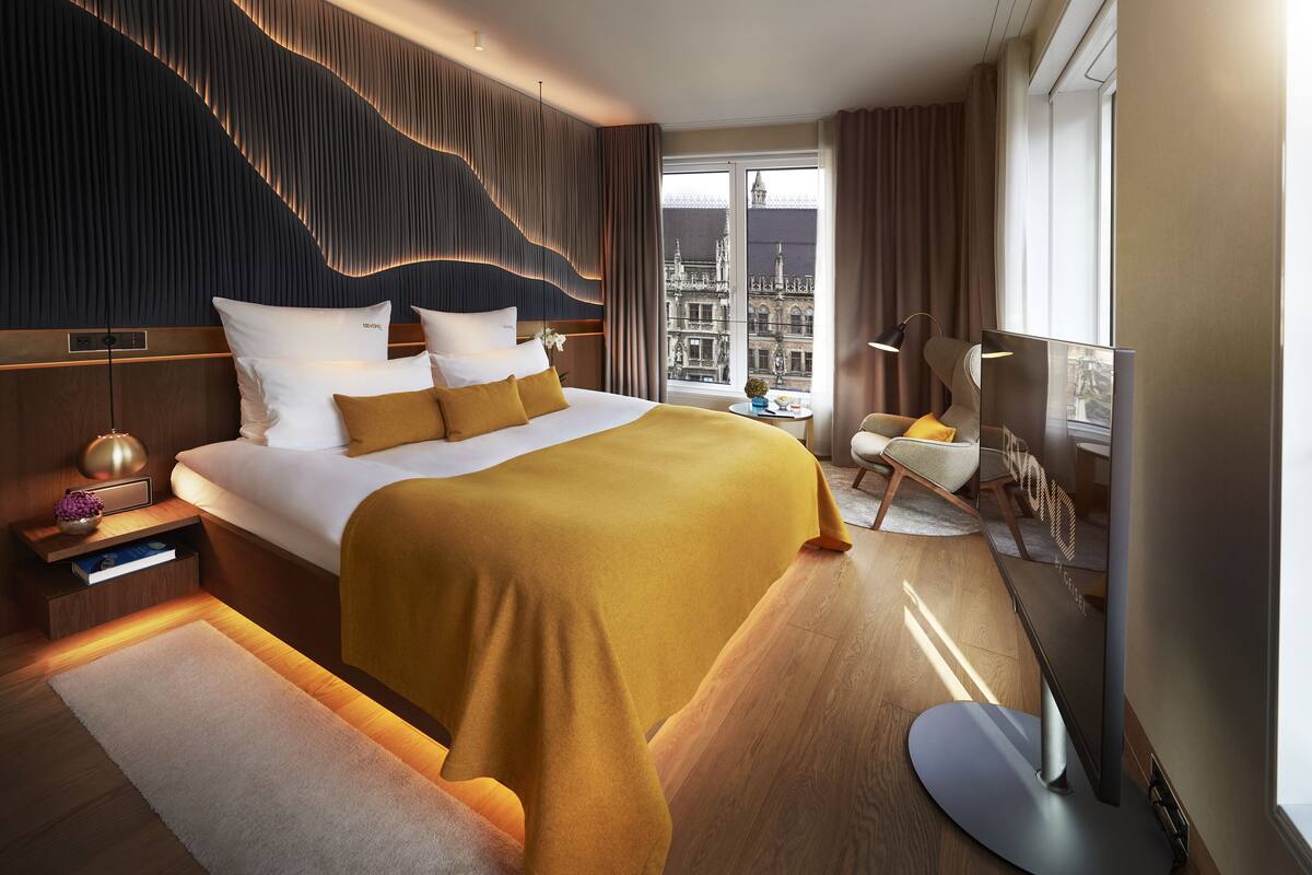 Et moderne hotelværelse med en stor seng, et fladskærms-TV, et natbord med lampe og udsigt til en historisk by gennem vinduet.