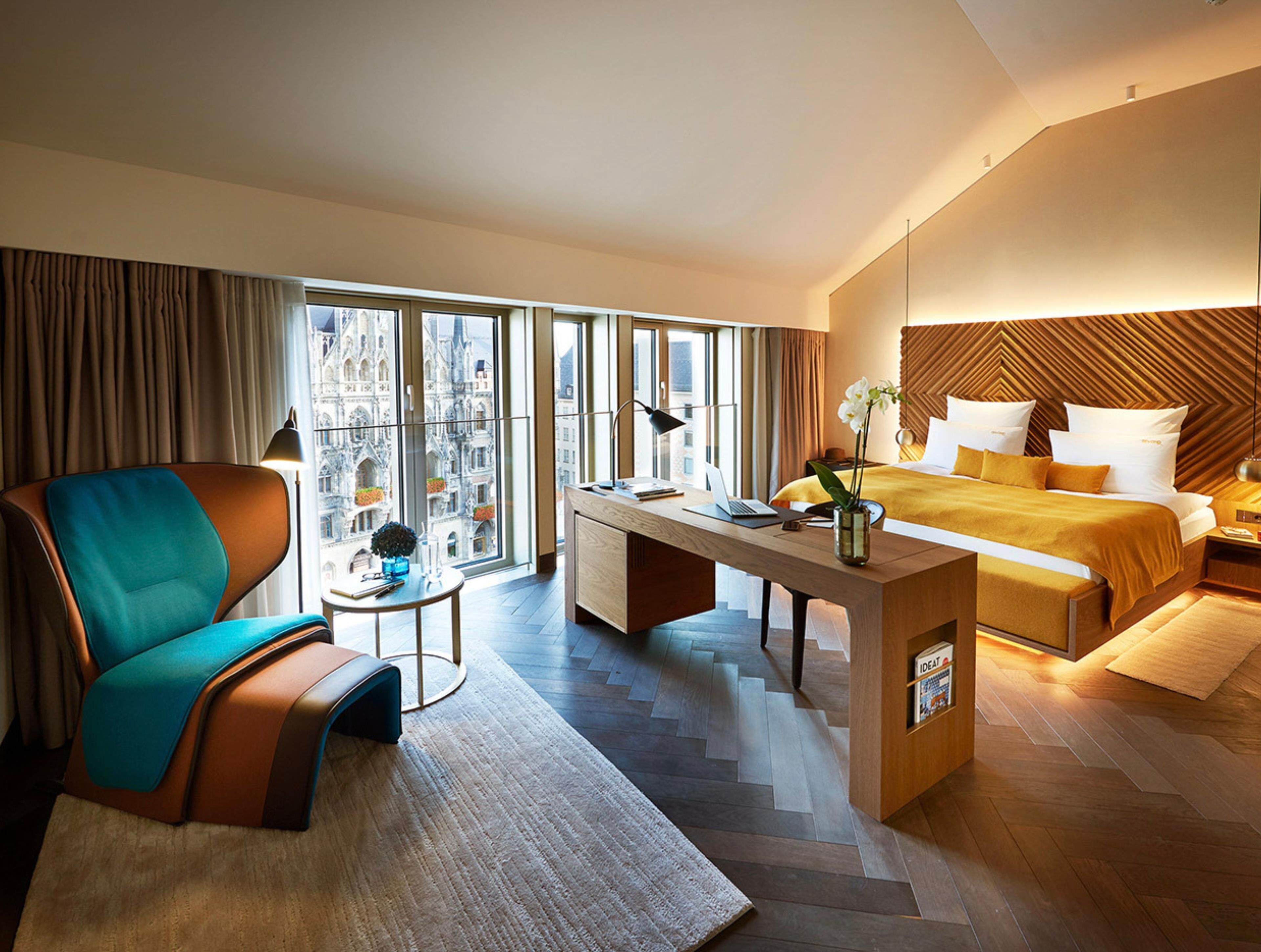 A modern hotel room with a large bed, a desk, a chair, and a view of a cityscape.