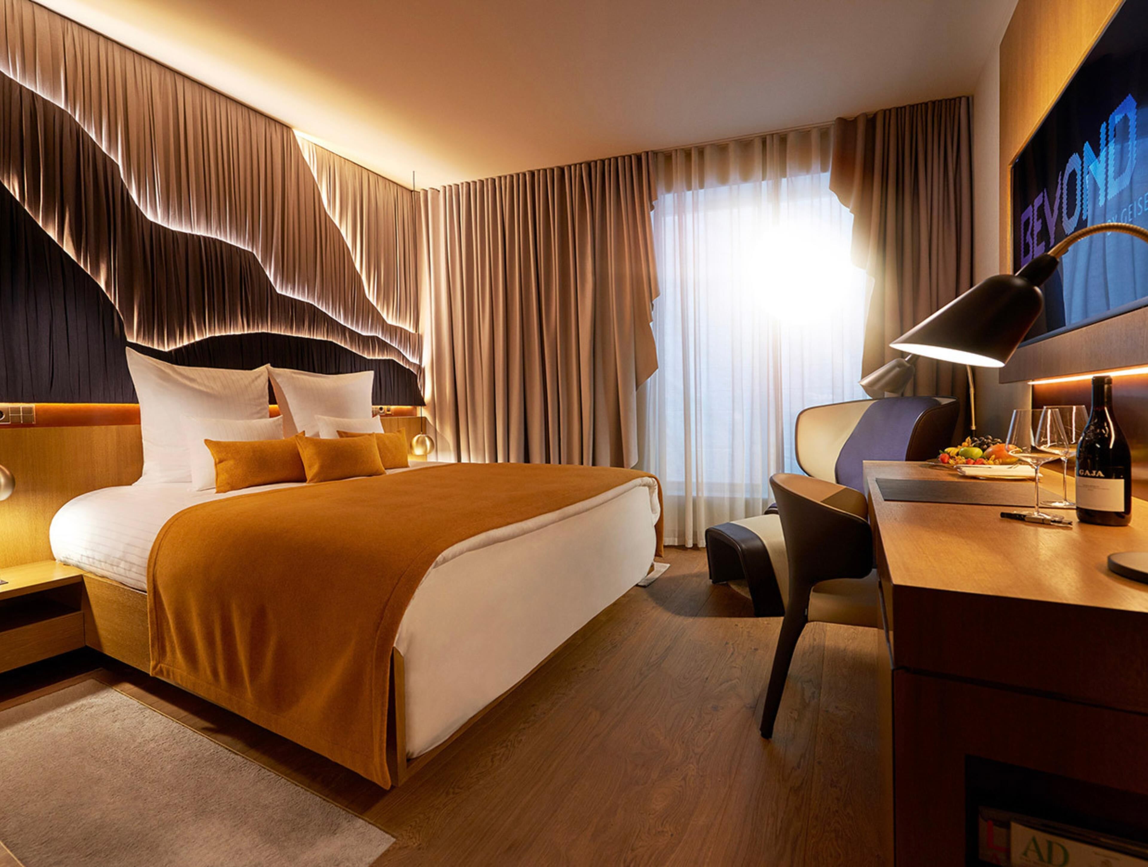 Ett hotellrum med en stor säng, ett skrivbord, en stol, en lampa och en minibar.