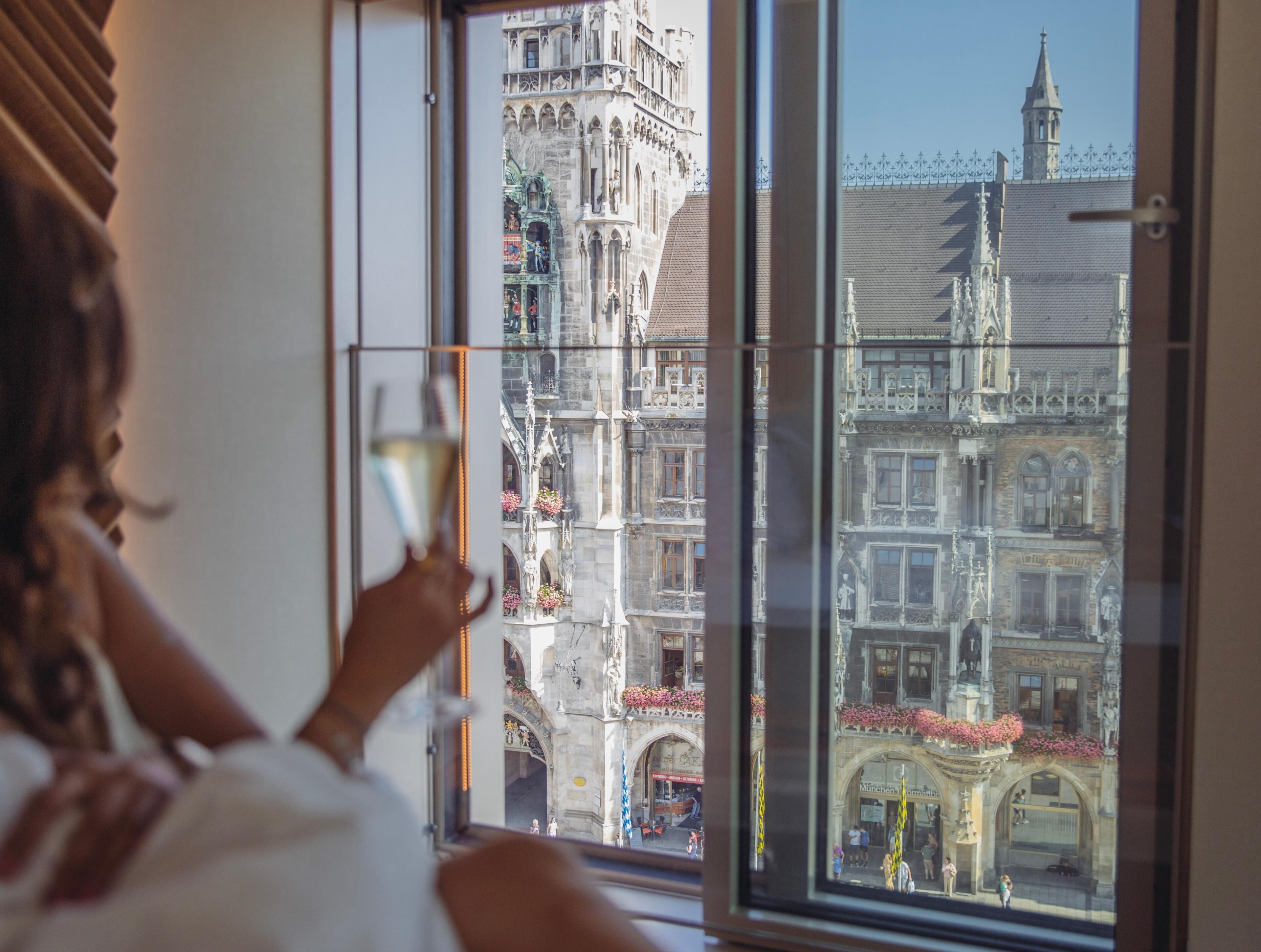 A person holding a glass, looking out of a window at a historic building with intricate architecture.