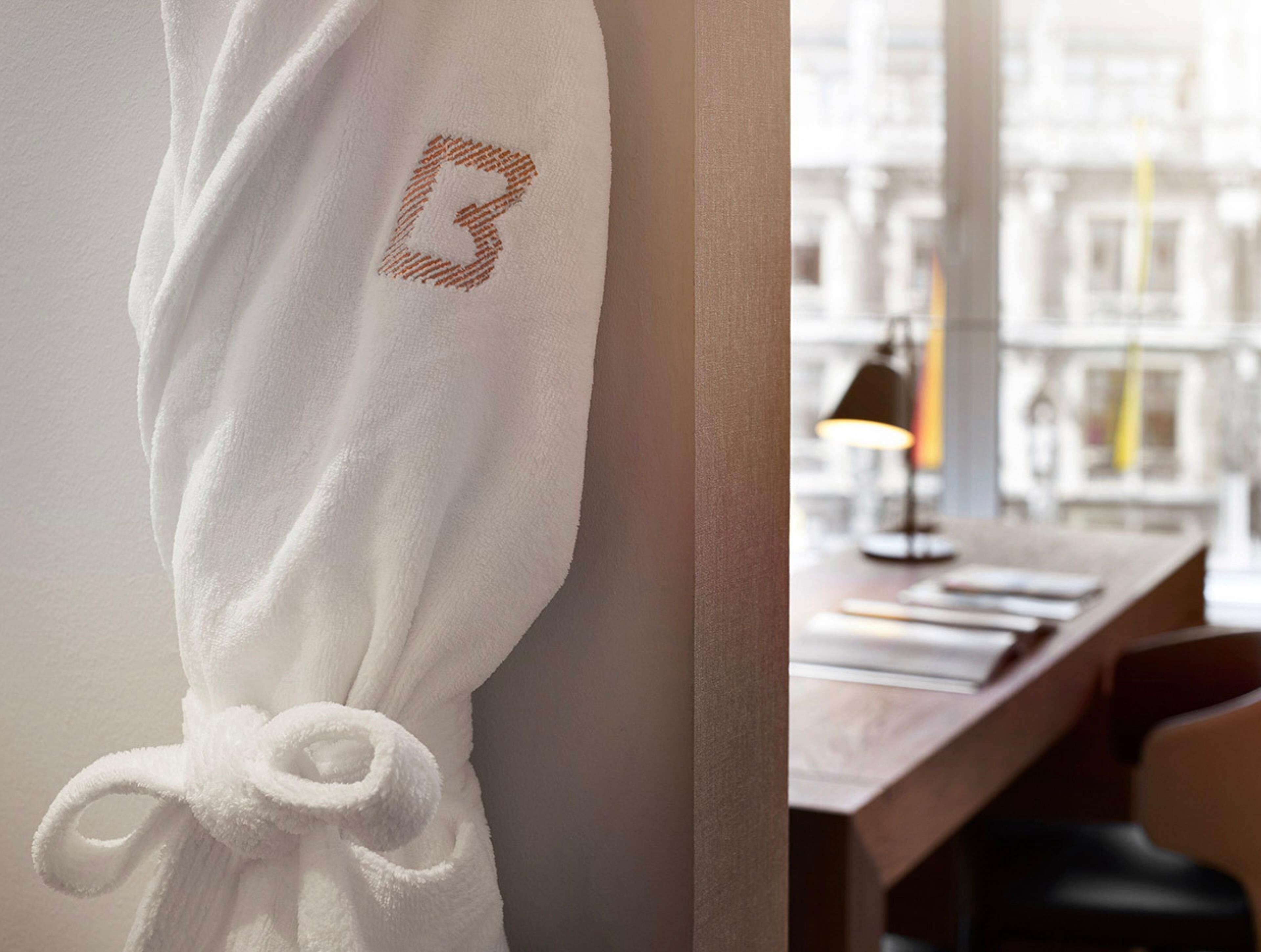A white robe with a monogrammed letter 'B' hanging on a wall, a wooden desk with a lamp, and a view of buildings through a window.
