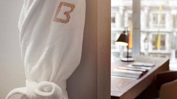 A white robe with a monogrammed letter 'B' hanging on a wall, a wooden desk with a lamp, and a view of buildings through a window.