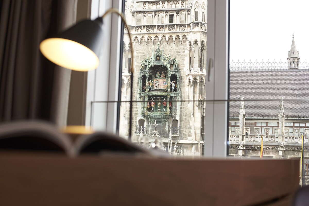Uitzicht door een raam met een lamp, waarop een historisch gebouw met gedetailleerde architectuur en een klokkentoren te zien is.