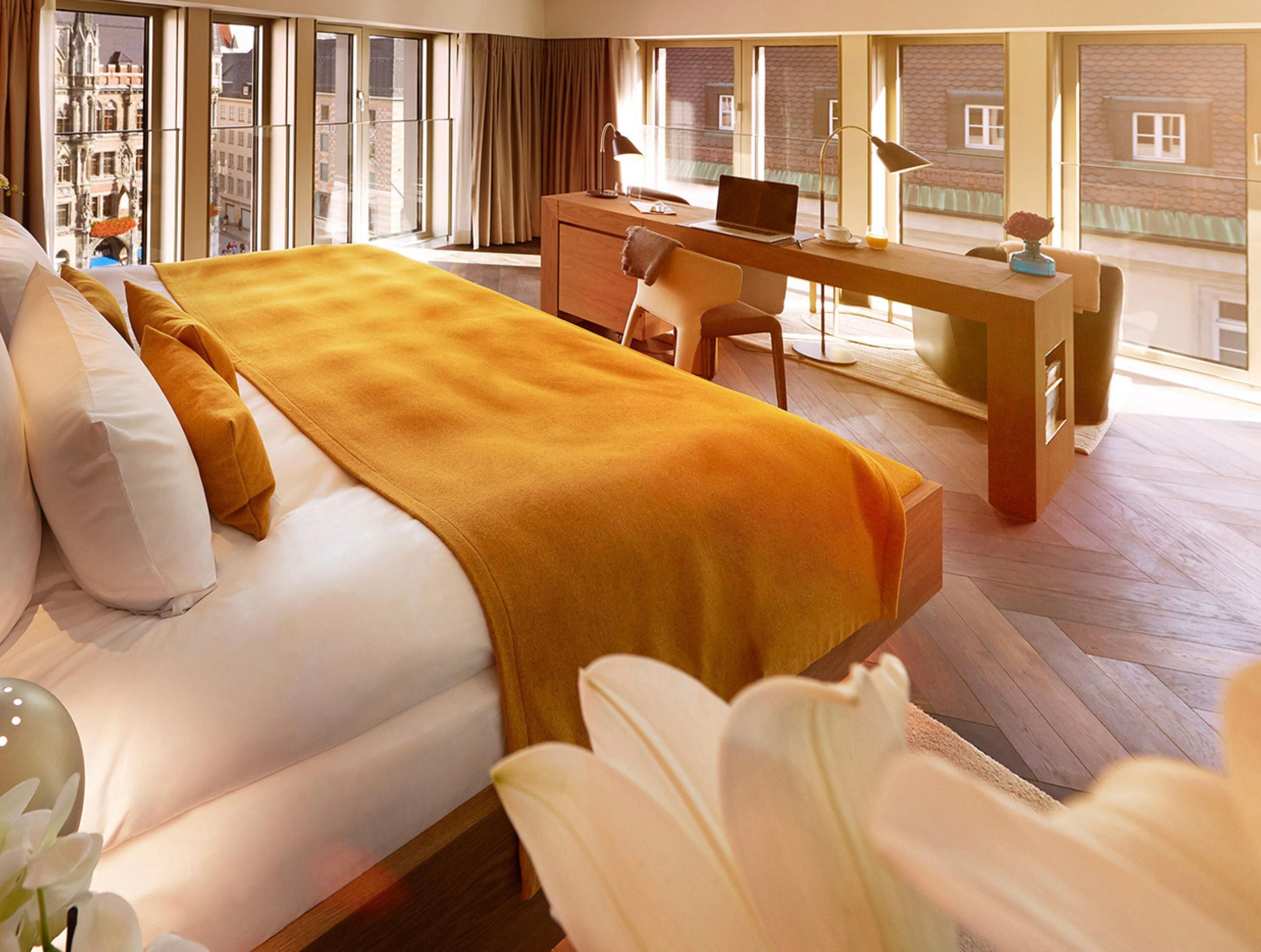 Habitación de hotel con cama, escritorio y vista a la ciudad a través de amplias ventanas.