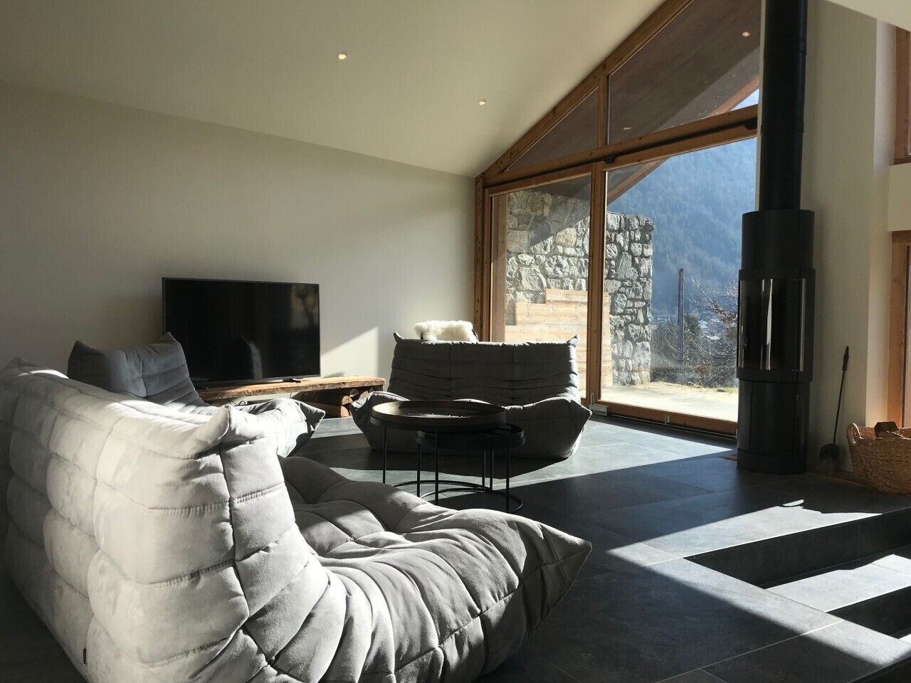Ein modernes Wohnzimmer mit einer Couch, einem Couchtisch und einem Fernseher. Ein großes Fenster bietet einen Ausblick ins Freie.