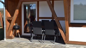 A wooden pergola with a glass door, two black chairs, and a barbecue grill. - HM- Holiday home 1 Deluxe Krombachtalsperre (Driedorf)