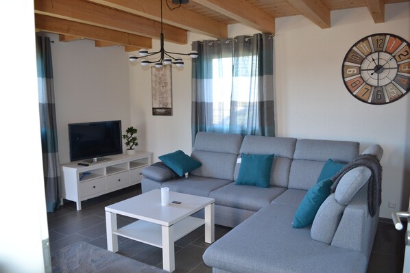 Una sala de estar con un sofá gris, una mesa de centro blanca, un televisor sobre un mueble blanco y un reloj de pared.