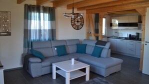 A modern living room with a grey sectional sofa, a white coffee table, and a kitchen area with white cabinets and a black countertop. - HM- Holiday home 1 Deluxe Krombachtalsperre (Driedorf)