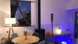 Una habitación acogedora con una mesa redonda de madera, un cuadro grande de un barco, un poste negro alto y una roca decorativa iluminada.
