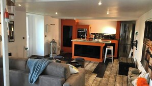 Una sala de estar moderna con un sofá gris, una mesita y una cocina con una isla central, un refrigerador y una estufa.