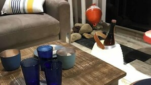 Una sala de estar con un sofá gris, una mesa de centro de madera con cristalería azul y gris, un jarrón rojo y una botella de champán.