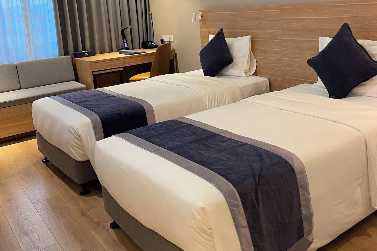 A hotel room with two beds, a desk, a chair, a window with curtains, and a painting on the wall.