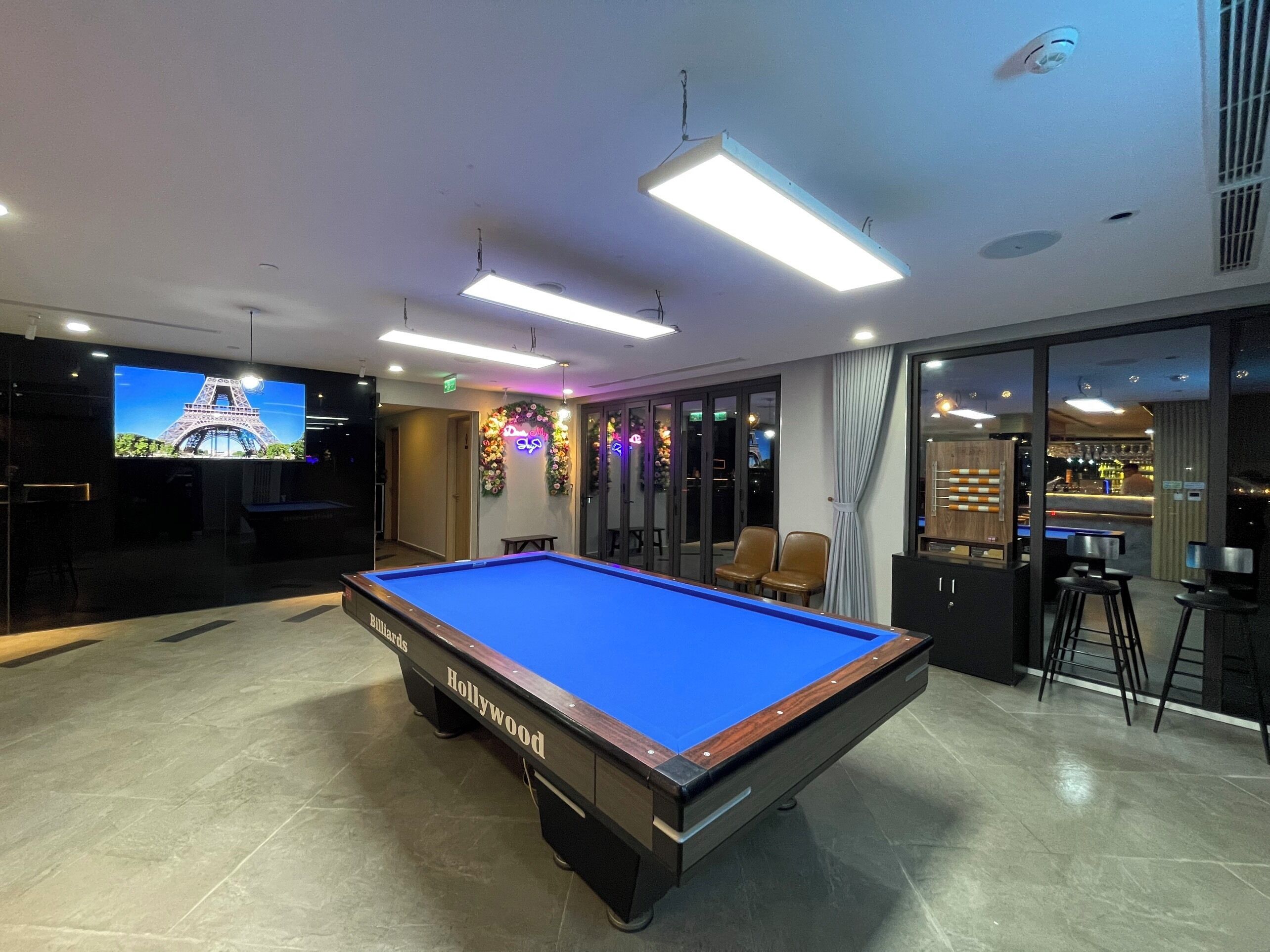 Une table de billard portant l’inscription « Hollywood » se trouve dans une pièce dotée d’un grand écran sur lequel est projetée la Tour Eiffel, ainsi que d’un bar et d’un espace détente avec des fauteuils.