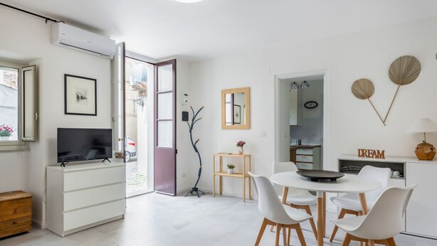 Ein moderner Wohnraum mit Flachbildfernseher, einem Essbereich mit weißen Stühlen, einer Küche mit weißem Schrank und einer Tür zu einem Außenbereich.