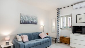 Ein modernes Wohnzimmer mit einem blauen Sofa, einem weißen Beistelltisch, einer Stehlampe, einem Fernseher und einem Gemälde an der Wand.