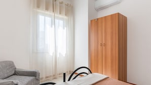 A bedroom with a bed, a grey sofa, a wooden wardrobe, and a window with sheer curtains. - GuestHost - Cozy 60 sqm apartment, for 4 people, with two bedroom, a terrace and a balcony, located on the 4th floor of a building with no lift. The property is located in a strategic position in Giardini Naxos, a seaside area near Taormina, less than 10 (Giardini Naxos)