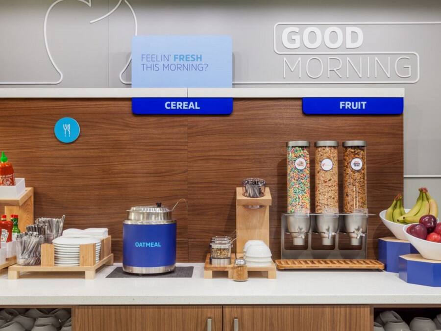 A breakfast bar with cereal and fruit options, a coffee machine, and condiments.