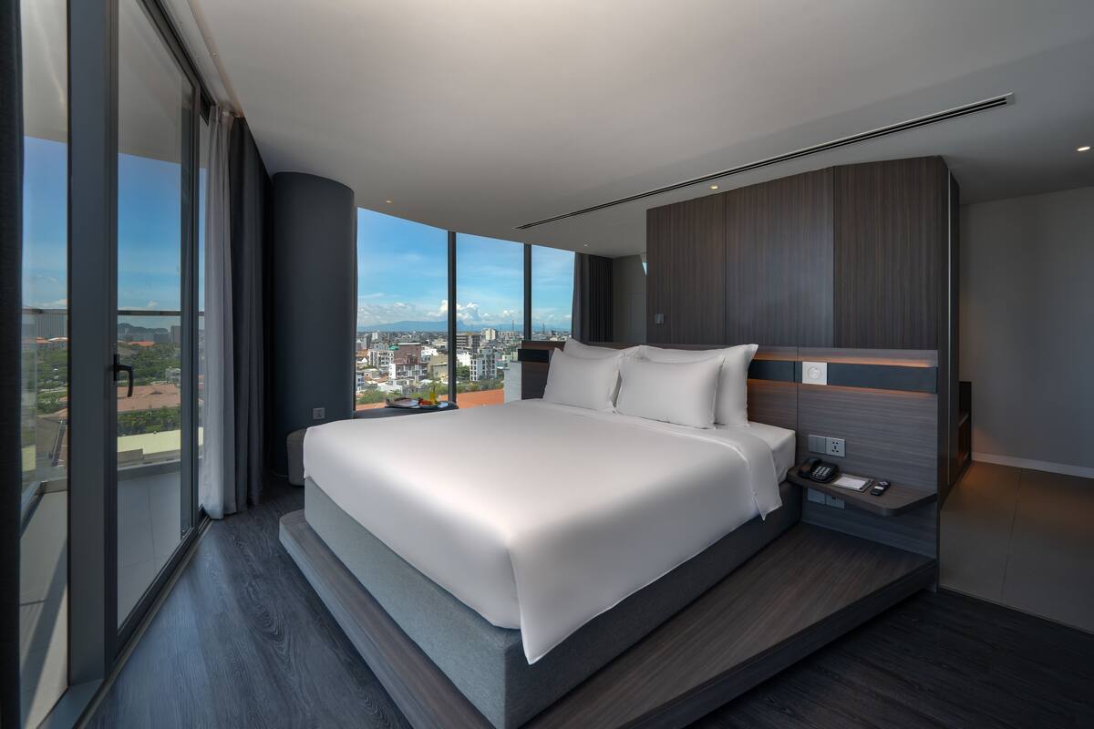 A modern bedroom with a large bed, a bedside table with a phone, and a city view through large windows.
