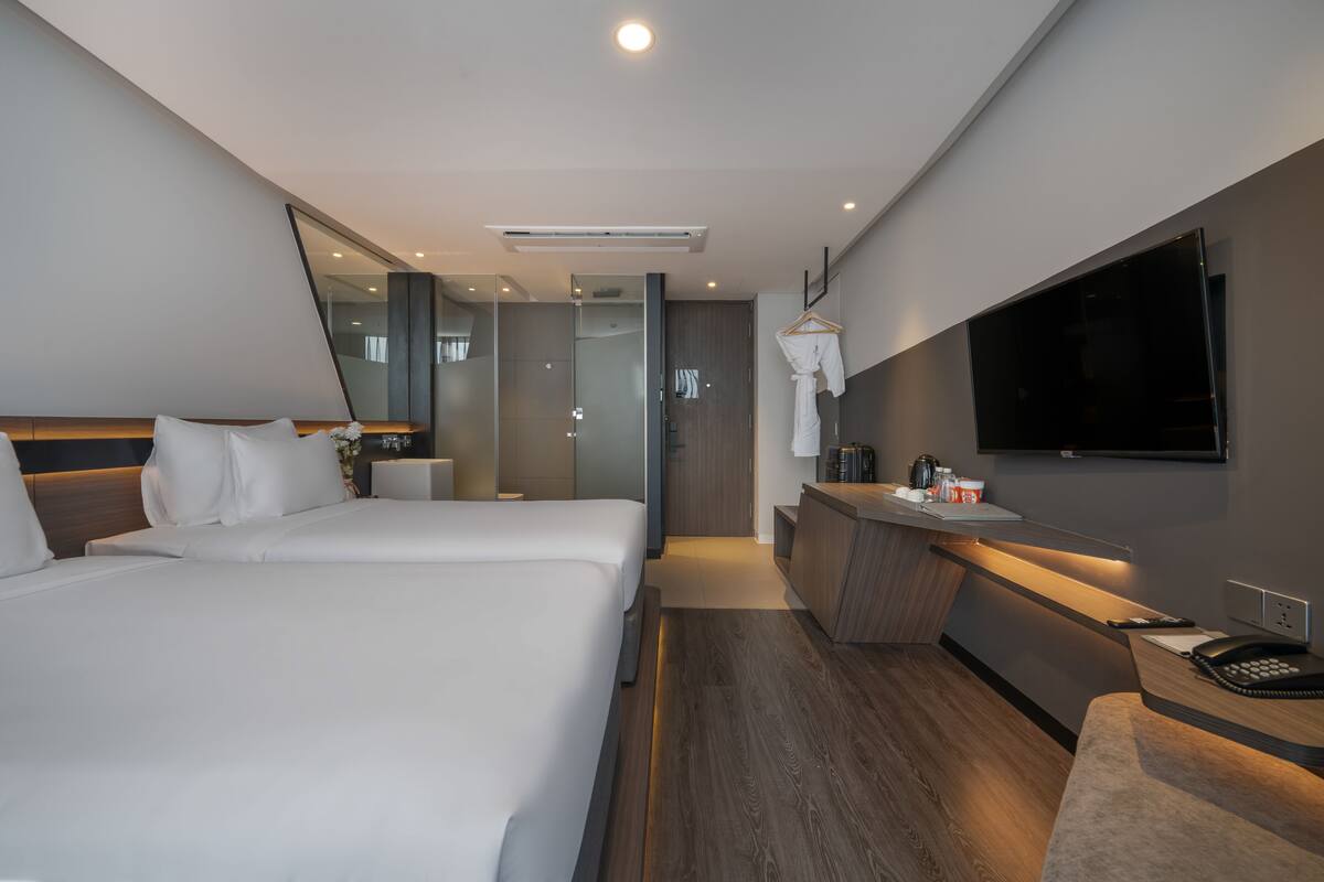 A modern hotel room with two beds, a desk, a mirror, a TV, and a shower area.
