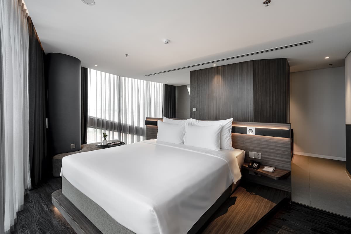 A modern hotel room with a large bed, a bedside table with a lamp, and a view of the city through large windows.