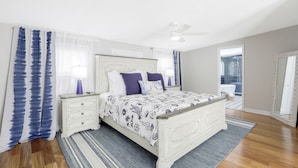A bedroom with a white bed, a nightstand, a ceiling fan, and a view of the bathroom.