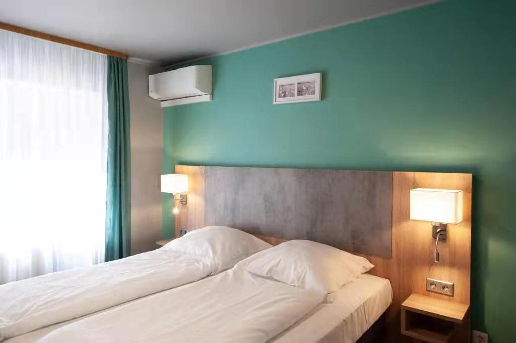 A hotel room with a bed, two bedside lamps, an air conditioning unit, and a framed picture on the wall.