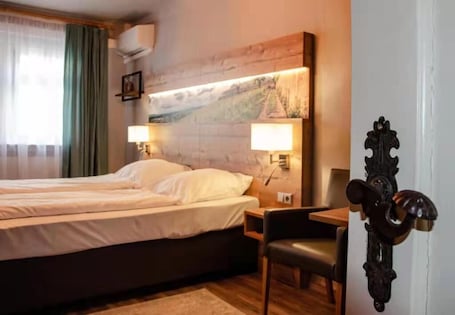 A hotel room with a double bed, two bedside lamps, a wooden headboard, a small table, a chair, and a decorative door handle.