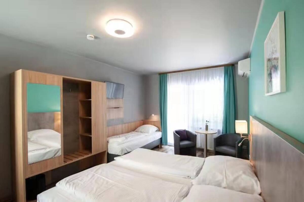 A hotel room with two single beds, a wooden wardrobe, a small table with a chair, and a window with curtains.