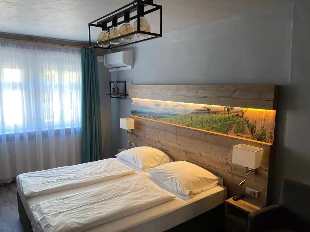 A hotel room with a large bed, a headboard with a landscape painting, a bedside table with a lamp, and a window with curtains.