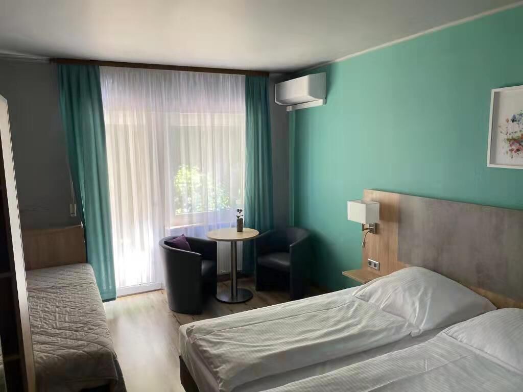 A hotel room with two beds, a small table, a chair, a window with curtains, and a wall-mounted air conditioner.