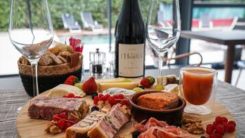 A wooden board with various cheeses, meats, and fruits, accompanied by wine glasses and a bottle of wine, set on a table with a view of a pool and lounge chairs.