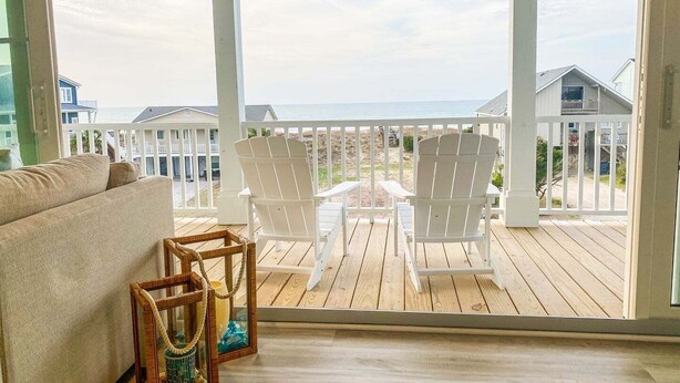Een houten terras met een witte veranda, twee witte Adirondack-stoelen, een beige bank en een rieten lantaarn.