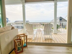 Une terrasse en bois avec un porche blanc, deux fauteuils Adirondack blancs, un canapé beige et une lanterne en osier.