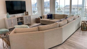 Een woonkamer met een hoekbank, een flatscreen televisie, een plafondventilator en grote ramen met uitzicht op gebouwen.
