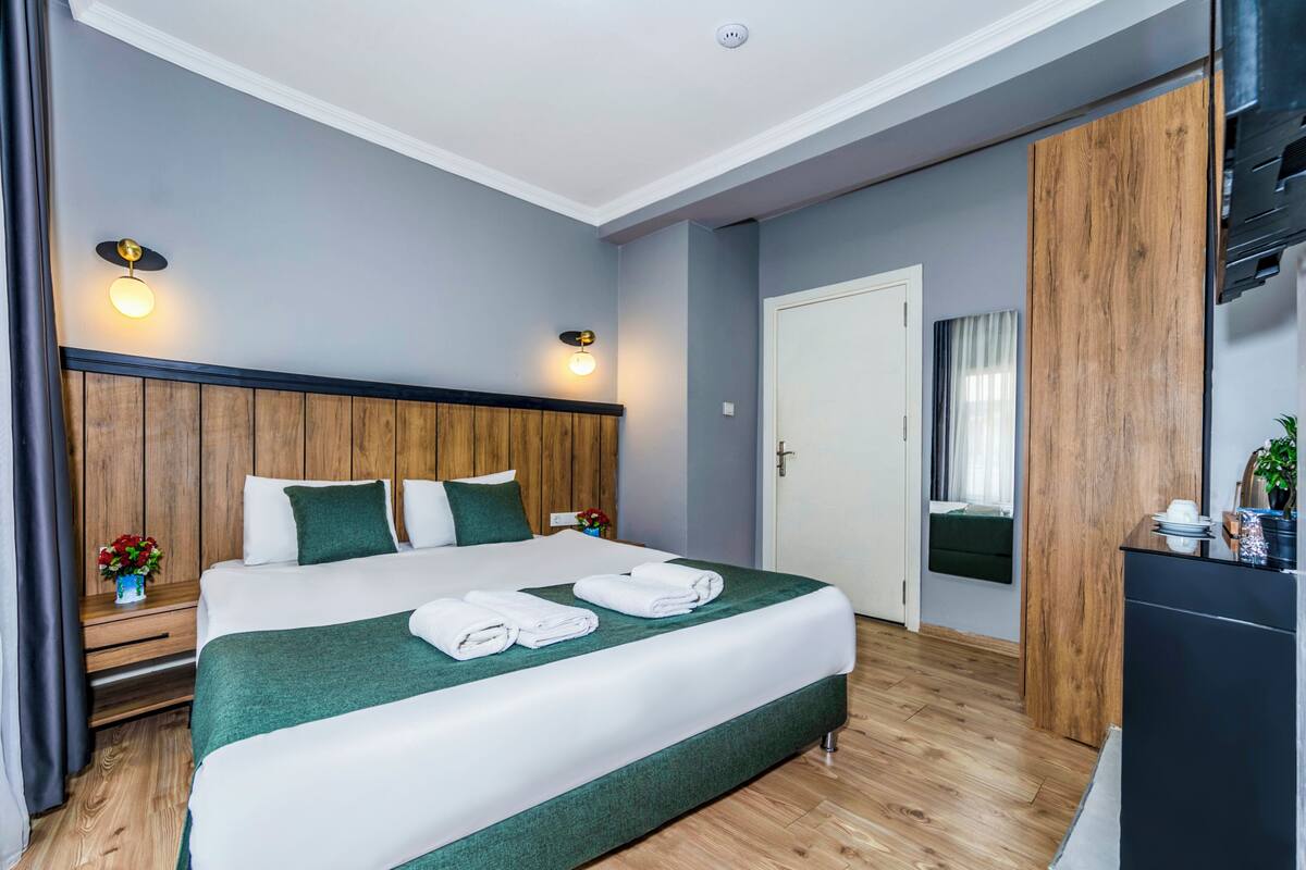 A hotel room with a wooden headboard, a bed with green bedding, two bedside lamps, a wooden floor, a door, and a small table with a plant.