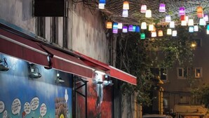 Un cafè al costat del carrer amb seients a l'aire lliure, un mural a la paret i llums penjants de colors.