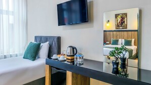 Habitació d'hotel amb un llit, una televisió penjada a la paret, una taula de nit amb una paperera, tasses i una planta.