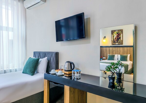 A hotel room with a bed, a TV mounted on the wall, a bedside table with a kettle, cups, and a plant.