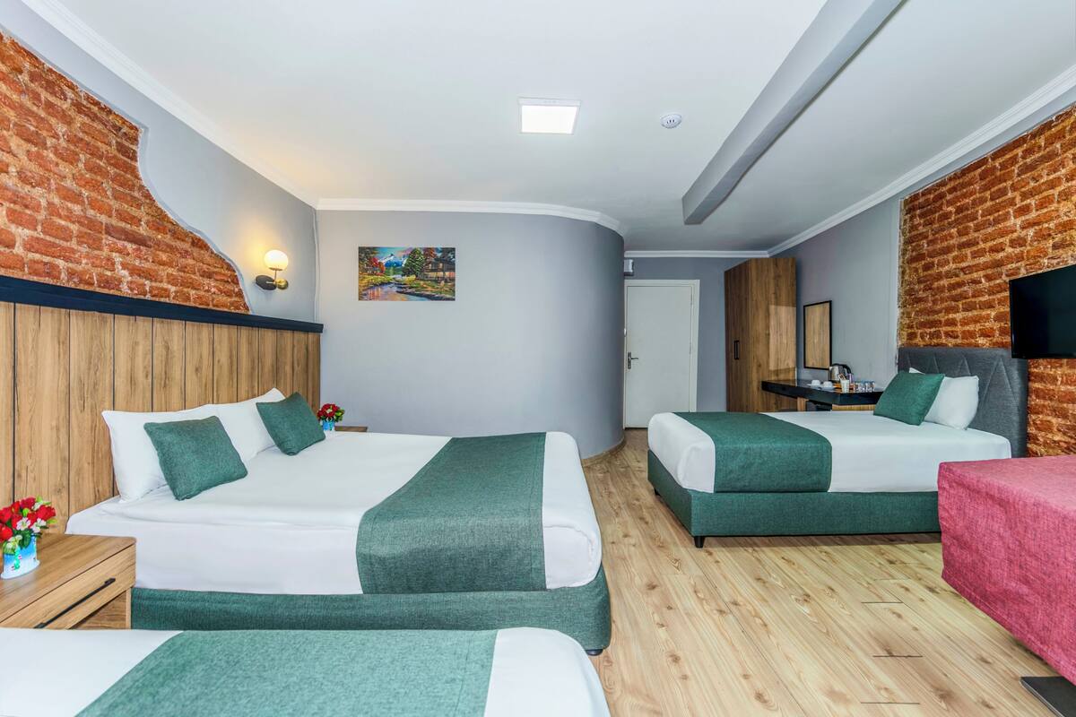 A hotel room with two beds, a wooden floor, a brick accent wall, a painting, and a small desk with a mirror.