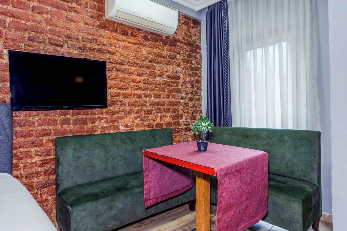 A room with a brick wall, a flat-screen TV mounted above a green sofa, a small table with a red tablecloth, and a potted plant.
