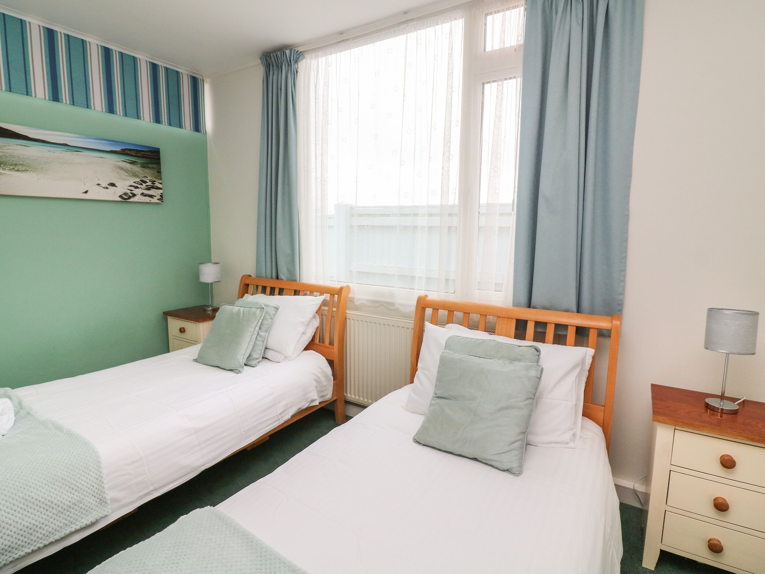 A twin room with two single beds, a nightstand with a lamp, a dresser, and a window with curtains.