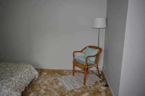 A small, sparsely furnished room with a single bed, a wicker chair, a standing lamp, and a patterned rug.