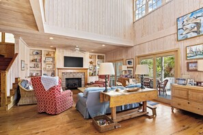 A spacious living room with a fireplace, comfortable seating, a television, and a large window with a view of greenery.