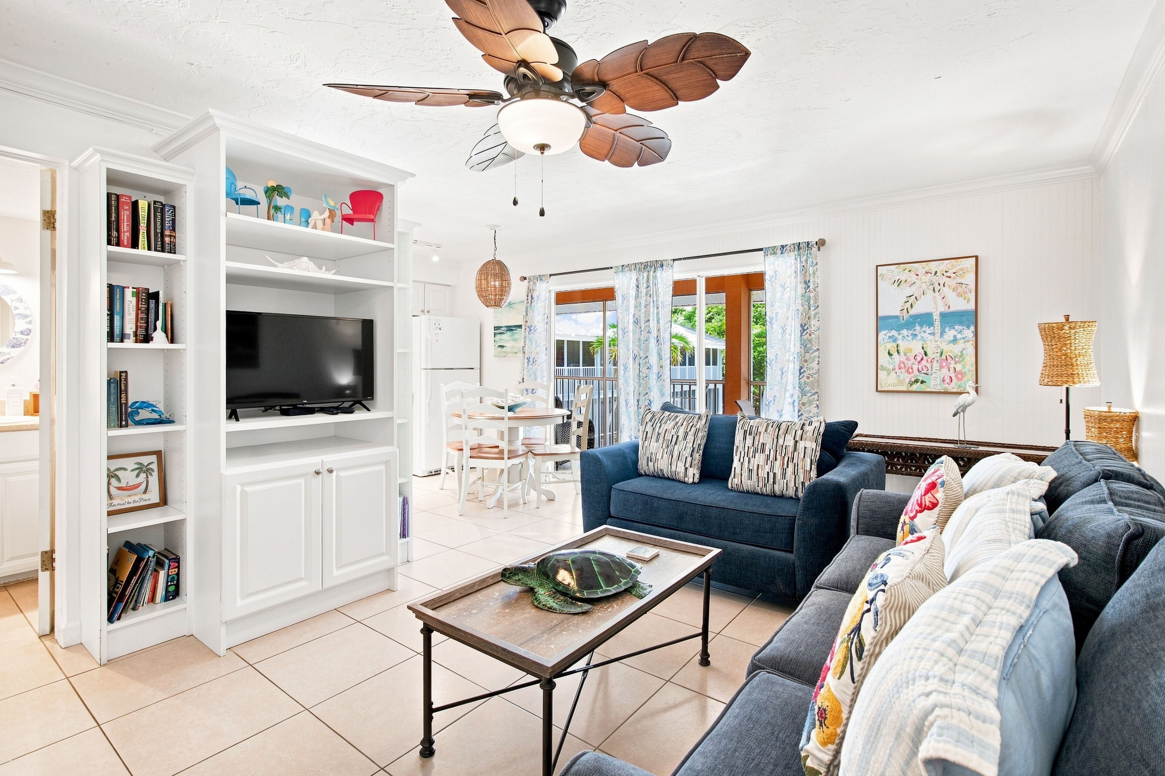 A living room with a TV, white cabinets, a blue sofa, a ceiling fan, and a view of the outdoors.