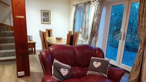 A red leather armchair with heart-shaped pillows in a living room with a dining area, a staircase, and large windows. - Inviting 3-bed Cottage Close to Pwllheli (Pwllheli)