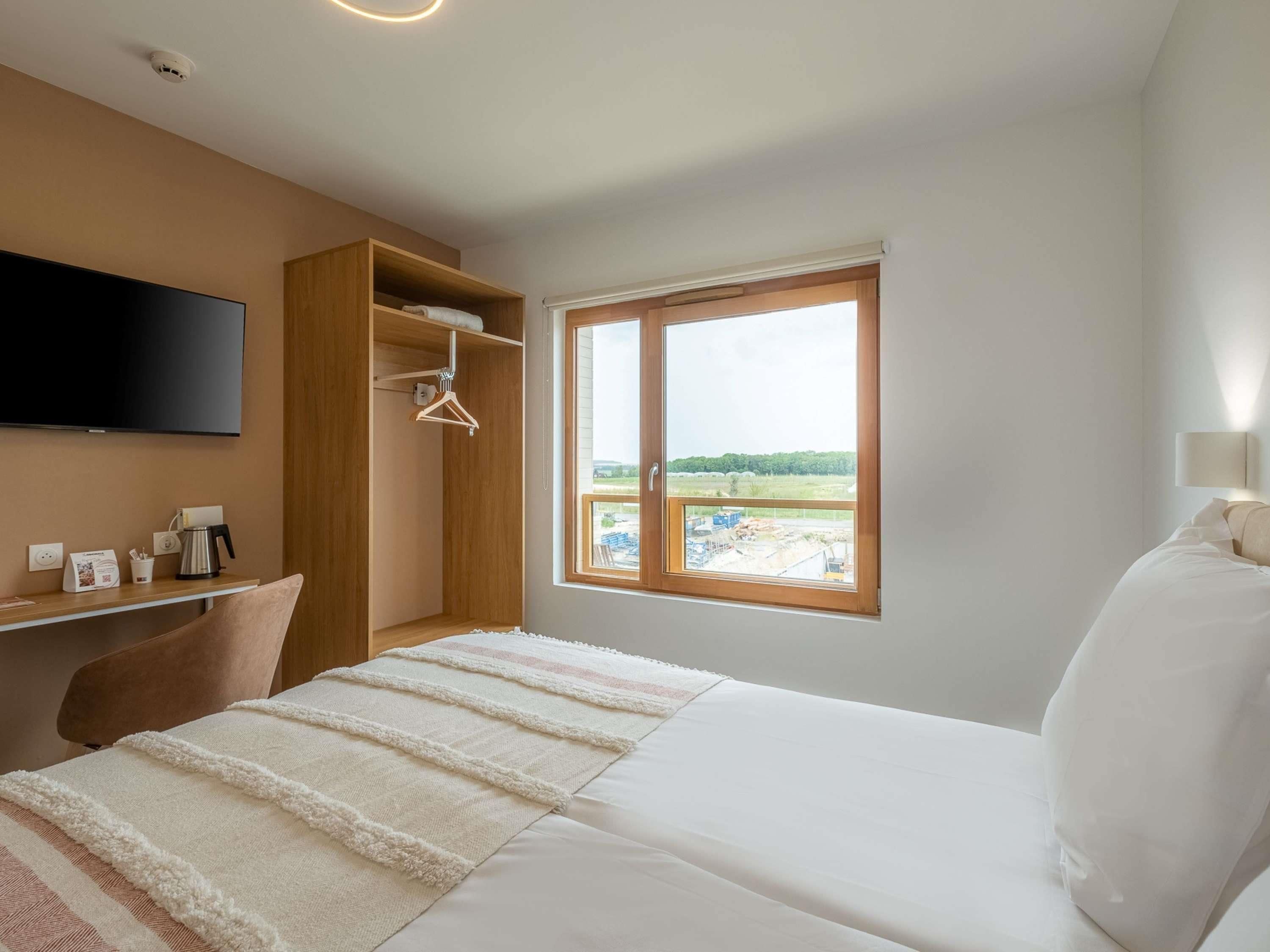 A hotel room with a bed, a TV, a desk with a coffee maker, a wardrobe, and a window with a view.