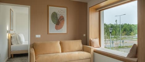 A modern room with a beige sofa, a large window with a view of a park, and a framed artwork on the wall.
